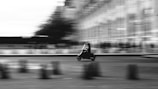 A motorbike is in motion, captured with a blurred background that suggests speed and movement. The image is in black and white, adding a classic and dynamic feel. The rider is silhouetted, emphasizing the sense of movement against the urban backdrop.