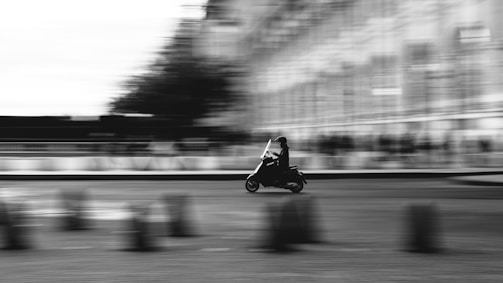 A motorbike is in motion, captured with a blurred background that suggests speed and movement. The image is in black and white, adding a classic and dynamic feel. The rider is silhouetted, emphasizing the sense of movement against the urban backdrop.