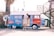 A man is standing at a blue and red retro-style van that has been converted into a coffee stand. The side of the van is open, revealing a coffee menu and the interior workspace. There is a palm tree to the left and a metal structure with stairs to the right. The background includes some fencing and outdoor seating equipment.