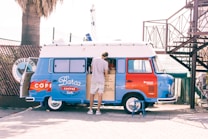A man is standing at a blue and red retro-style van that has been converted into a coffee stand. The side of the van is open, revealing a coffee menu and the interior workspace. There is a palm tree to the left and a metal structure with stairs to the right. The background includes some fencing and outdoor seating equipment.