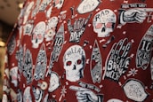 The image features a fabric with a bold, graphic pattern of white skulls wearing red 3D glasses, surrounded by various surfing-related elements like hands with surf-related text, surfboards, and text bubbles. The background is a deep red, creating a striking contrast with the white and black illustrations. The design includes phrases like 'CHILLAX,' 'RAD TO THE BONE,' and 'RIDE THIS WAY,' adding a playful, rebellious vibe.
