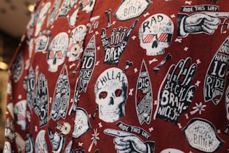 The image features a fabric with a bold, graphic pattern of white skulls wearing red 3D glasses, surrounded by various surfing-related elements like hands with surf-related text, surfboards, and text bubbles. The background is a deep red, creating a striking contrast with the white and black illustrations. The design includes phrases like 'CHILLAX,' 'RAD TO THE BONE,' and 'RIDE THIS WAY,' adding a playful, rebellious vibe.