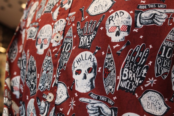 The image features a fabric with a bold, graphic pattern of white skulls wearing red 3D glasses, surrounded by various surfing-related elements like hands with surf-related text, surfboards, and text bubbles. The background is a deep red, creating a striking contrast with the white and black illustrations. The design includes phrases like 'CHILLAX,' 'RAD TO THE BONE,' and 'RIDE THIS WAY,' adding a playful, rebellious vibe.