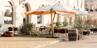 An outdoor dining area is set outside a stylish building with arched windows and elegant wall sconces. Large white and orange umbrellas provide shade over the seating area, which includes several tables and chairs. Greenery like potted plants and small trees add to the relaxing atmosphere. The ground is paved with warm-toned stones, and a wooden barrel is used as decoration.