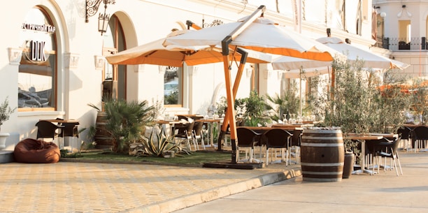 An outdoor dining area is set outside a stylish building with arched windows and elegant wall sconces. Large white and orange umbrellas provide shade over the seating area, which includes several tables and chairs. Greenery like potted plants and small trees add to the relaxing atmosphere. The ground is paved with warm-toned stones, and a wooden barrel is used as decoration.