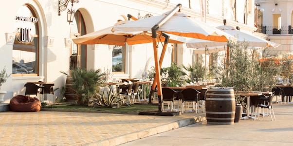 An outdoor dining area is set outside a stylish building with arched windows and elegant wall sconces. Large white and orange umbrellas provide shade over the seating area, which includes several tables and chairs. Greenery like potted plants and small trees add to the relaxing atmosphere. The ground is paved with warm-toned stones, and a wooden barrel is used as decoration.
