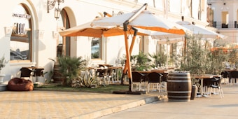 An outdoor dining area is set outside a stylish building with arched windows and elegant wall sconces. Large white and orange umbrellas provide shade over the seating area, which includes several tables and chairs. Greenery like potted plants and small trees add to the relaxing atmosphere. The ground is paved with warm-toned stones, and a wooden barrel is used as decoration.
