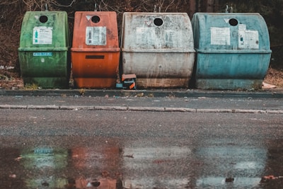 Four recycling bins in different colors, green, orange, and two shades of blue, are placed along a paved street. The bins are situated in front of a wooded area with fallen leaves around. The ground is wet, creating reflections of the bins in a shallow puddle.