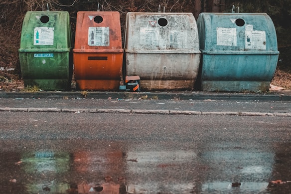 Four recycling bins in different colors, green, orange, and two shades of blue, are placed along a paved street. The bins are situated in front of a wooded area with fallen leaves around. The ground is wet, creating reflections of the bins in a shallow puddle.