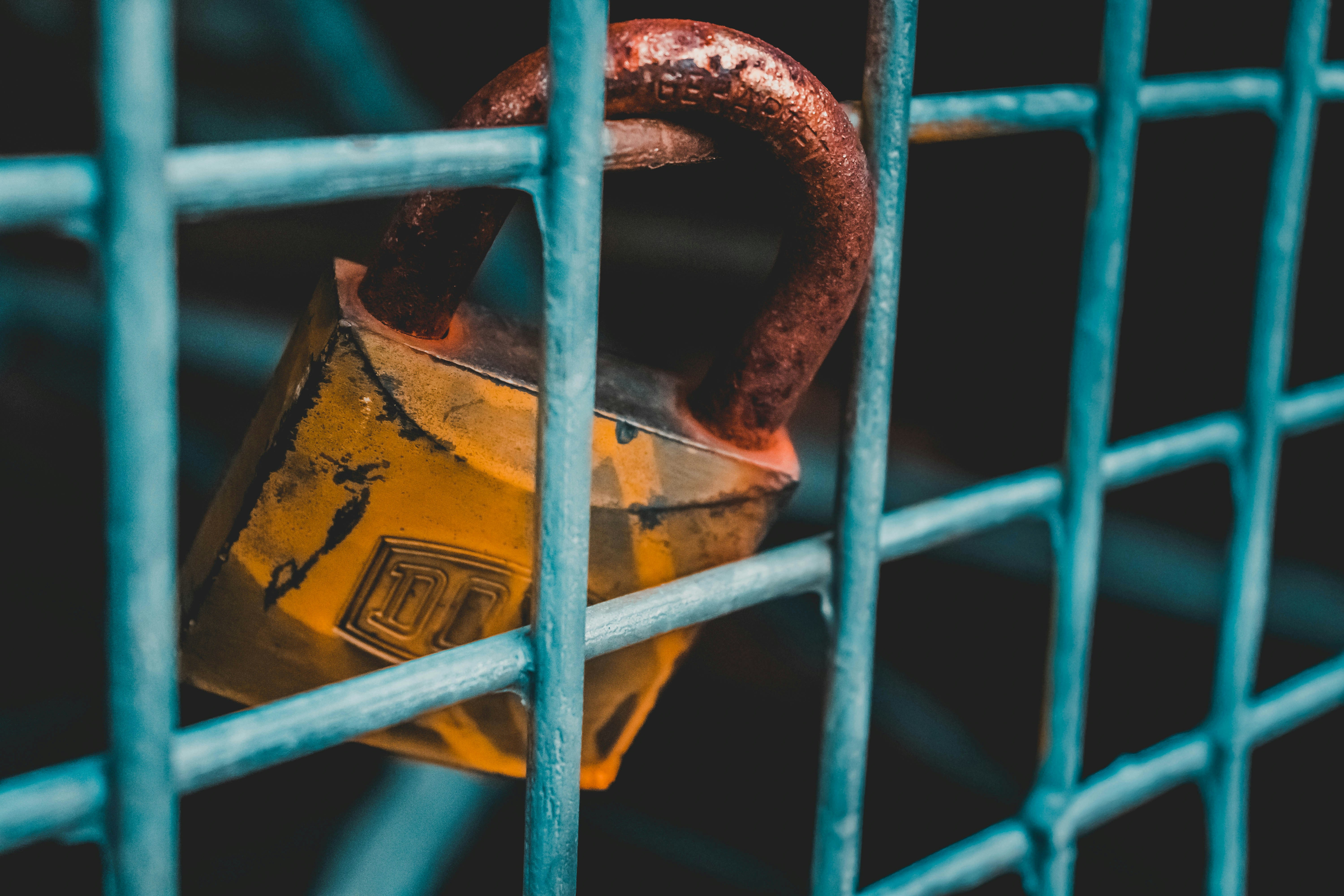 Close-up of a weathered yellow padlock with a rusted shackle securing a blue metal fence, symbolizing outdated security or resistance to change.