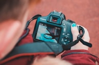A person holds a Canon camera, focusing on an image of a basketball hoop against a blue sky with clouds. The individual's nails are painted blue, matching the sky on the camera screen.