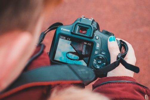 A person holds a Canon camera, focusing on an image of a basketball hoop against a blue sky with clouds. The individual's nails are painted blue, matching the sky on the camera screen.