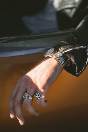A close-up of a person's hand adorned with multiple rings, showcasing intricate designs with turquoise stones. The hand rests casually against a surface, and the person is wearing a leather jacket with a visible zipper. A watch with a decorative band is also visible on the wrist.