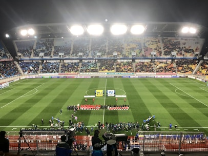 A large soccer stadium with bright overhead lights and a well-maintained green field. Teams are lined up in the center alongside officials, and there is a group gathered nearby, possibly for a pre-match ceremony. The stadium is filled with spectators wearing colorful clothing, and banners and advertisements are visible along the edges of the field. Cameras and officials are positioned on the sidelines to capture the event.