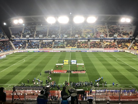 A large soccer stadium with bright overhead lights and a well-maintained green field. Teams are lined up in the center alongside officials, and there is a group gathered nearby, possibly for a pre-match ceremony. The stadium is filled with spectators wearing colorful clothing, and banners and advertisements are visible along the edges of the field. Cameras and officials are positioned on the sidelines to capture the event.