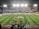 A large soccer stadium with bright overhead lights and a well-maintained green field. Teams are lined up in the center alongside officials, and there is a group gathered nearby, possibly for a pre-match ceremony. The stadium is filled with spectators wearing colorful clothing, and banners and advertisements are visible along the edges of the field. Cameras and officials are positioned on the sidelines to capture the event.
