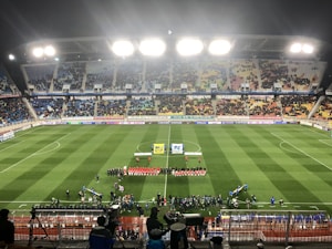 A large soccer stadium with bright overhead lights and a well-maintained green field. Teams are lined up in the center alongside officials, and there is a group gathered nearby, possibly for a pre-match ceremony. The stadium is filled with spectators wearing colorful clothing, and banners and advertisements are visible along the edges of the field. Cameras and officials are positioned on the sidelines to capture the event.