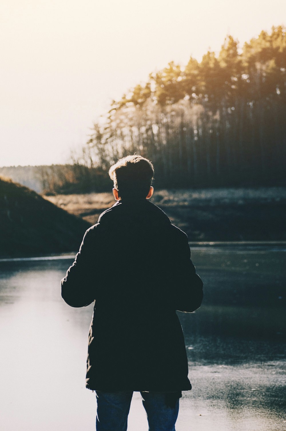 man standing infront of lake and trees during daytime