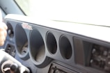 The interior section of a vehicle dashboard is visible, showcasing several circular openings where dials or gauges would typically be located. To the left, part of a steering wheel or similar control apparatus is visible. The dashboard appears to lack instruments, suggesting a deconstructed or unfinished vehicle.