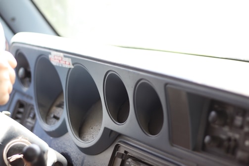 The interior section of a vehicle dashboard is visible, showcasing several circular openings where dials or gauges would typically be located. To the left, part of a steering wheel or similar control apparatus is visible. The dashboard appears to lack instruments, suggesting a deconstructed or unfinished vehicle.