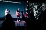 A dimly lit stage with musicians performing, featuring one person playing a guitar. There are silhouettes of people in the foreground with hands raised, and lyrics displayed on a screen in the background.