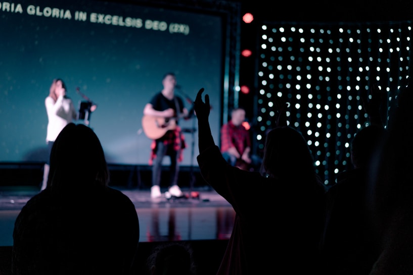 A dimly lit stage with musicians performing, featuring one person playing a guitar. There are silhouettes of people in the foreground with hands raised, and lyrics displayed on a screen in the background.