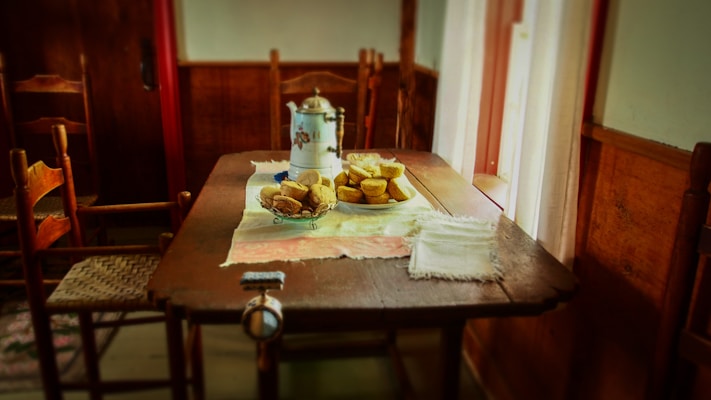 A rustic wooden table set in a cozy room with wooden chairs. On the table, there are pastries or bread rolls arranged on a plate and a metal coffee pot. A folded cloth napkin lies beside it, with soft lighting coming through a window with sheer curtains.