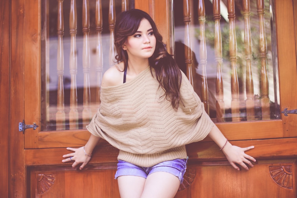 woman in brown one-shoulder top and blue denim shorts leaning on brown wooden door