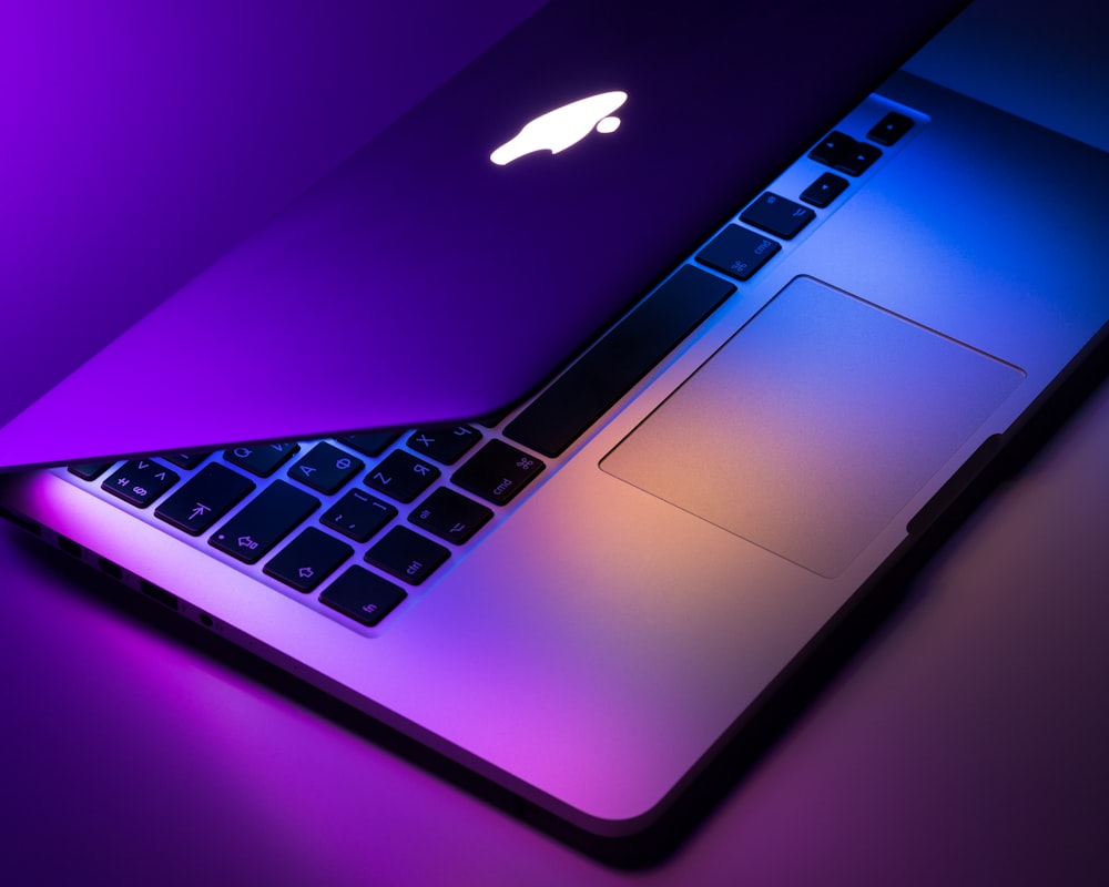 A refurbished Apple Macbook in a dark room with the screen half open illuminating the keyboard in multiple colours