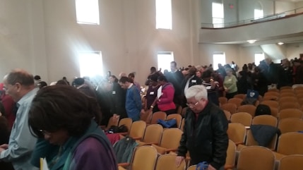 A large group of people standing in an auditorium. The room is well-lit with natural light streaming in from tall windows. The attendees appear engaged and are either looking down or focused on something in front of them. The seating consists of rows of beige chairs, and several individuals are wearing name tags.
