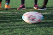 A rugby ball is positioned on a grassy field, with the text 'Get Into Rugby' printed on it. In the background, three pairs of feet wearing athletic shoes stand, suggesting players ready to engage in a rugby game.