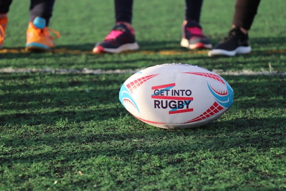 A rugby ball is positioned on a grassy field, with the text 'Get Into Rugby' printed on it. In the background, three pairs of feet wearing athletic shoes stand, suggesting players ready to engage in a rugby game.