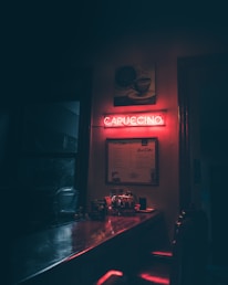 A dimly lit cafe interior features a glowing neon sign reading 'CAPUCCINO' in red. Below the sign, a menu hangs on the wall, and a counter is visible with various items like a coffee machine and bottles. The ambiance is cozy with shadows and warm lighting.