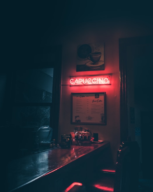 A dimly lit cafe interior features a glowing neon sign reading 'CAPUCCINO' in red. Below the sign, a menu hangs on the wall, and a counter is visible with various items like a coffee machine and bottles. The ambiance is cozy with shadows and warm lighting.