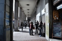 A group of people stands under a long, covered walkway with tall columns. Some are casually interacting, while others walk by. Various signs and graffiti are visible on the walls, and one chalkboard sign offers a promotion for hot chocolate and alfajor.