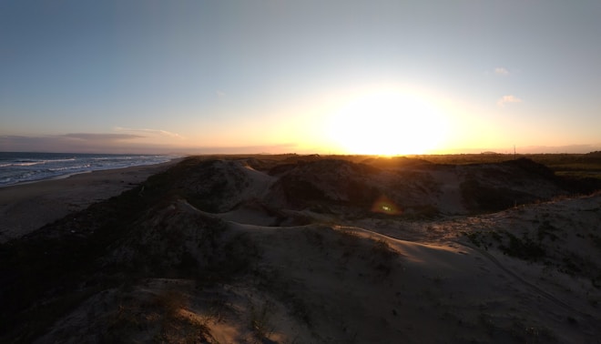 A serene sunset over sandy dunes with the sun casting a warm light across the landscape. The ocean is visible to the left with gentle waves, and the silhouette of the dunes creates a peaceful and expansive view.