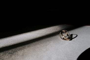 A pair of intertwined golden rings are placed on a textured surface, illuminated by a narrow beam of light against a dark background. The light emphasizes the metallic sheen of the rings and casts a soft shadow.