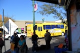 A busy urban street scene features a bright yellow bus labeled 'transborde' surrounded by several pedestrians. The background contains a commercial building with colorful signs, while trees and a lamppost are visible. The sunlight casts shadows, creating a vibrant and lively atmosphere.