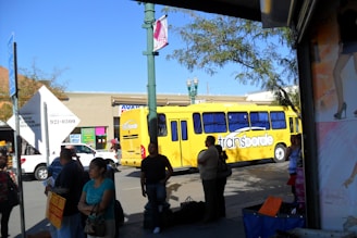 A busy urban street scene features a bright yellow bus labeled 'transborde' surrounded by several pedestrians. The background contains a commercial building with colorful signs, while trees and a lamppost are visible. The sunlight casts shadows, creating a vibrant and lively atmosphere.