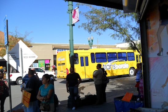 A busy urban street scene features a bright yellow bus labeled 'transborde' surrounded by several pedestrians. The background contains a commercial building with colorful signs, while trees and a lamppost are visible. The sunlight casts shadows, creating a vibrant and lively atmosphere.
