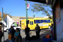 A busy urban street scene features a bright yellow bus labeled 'transborde' surrounded by several pedestrians. The background contains a commercial building with colorful signs, while trees and a lamppost are visible. The sunlight casts shadows, creating a vibrant and lively atmosphere.