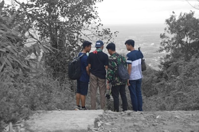 Four young men are standing together on a path surrounded by dense trees and vegetation. They appear to be looking at something in the distance. One of them is wearing a camouflage jacket, and two of them have backpacks. The surrounding environment seems to be a hilly or mountainous area.