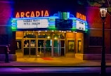 A vintage theater marquee illuminated at night, displaying movie titles 'Sausage Party' and 'Pete's Dragon.' The building features neon lights in vibrant colors, adding a nostalgic ambiance. A person stands near the entrance, checking their phone, while the interior lights glow warmly.