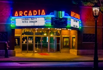 A vintage theater marquee illuminated at night, displaying movie titles 'Sausage Party' and 'Pete's Dragon.' The building features neon lights in vibrant colors, adding a nostalgic ambiance. A person stands near the entrance, checking their phone, while the interior lights glow warmly.
