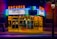 A vintage theater marquee illuminated at night, displaying movie titles 'Sausage Party' and 'Pete's Dragon.' The building features neon lights in vibrant colors, adding a nostalgic ambiance. A person stands near the entrance, checking their phone, while the interior lights glow warmly.
