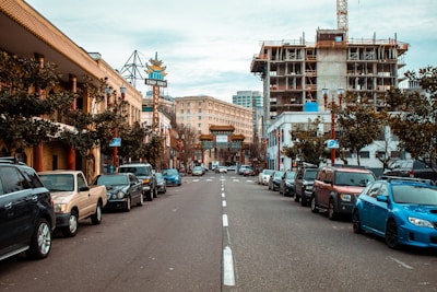 A street view of a city with cars parked on both sides. There are buildings on either side; one of the buildings to the right is under construction with scaffolding and cranes visible. The street leads to an ornate Chinatown gate in the distance. There are trees along the sidewalk and various signs, including one that reads 'Chop Suey.'