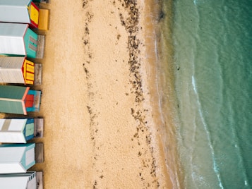 Aerial view of brightly colored beach huts lined up along a sandy shoreline, with gentle waves lapping against the shore and patches of seaweed scattered on the sand.