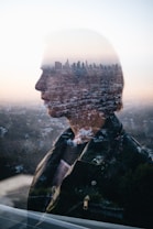 A double-exposure photograph combining a side profile of a person with an urban cityscape in the background. The image blends the silhouette and features of the person seamlessly with the tall city buildings and landscape, creating a surreal and artistic effect.