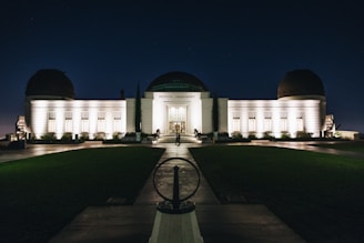 An illuminated observatory building stands against a dark night sky, its lights casting a warm glow on the white facade. The structure features a central dome flanked by two smaller domes, and a symmetrical arrangement of windows. A pathway leads up to the entrance, with neatly trimmed lawns on either side.