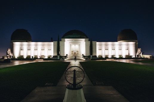 An illuminated observatory building stands against a dark night sky, its lights casting a warm glow on the white facade. The structure features a central dome flanked by two smaller domes, and a symmetrical arrangement of windows. A pathway leads up to the entrance, with neatly trimmed lawns on either side.